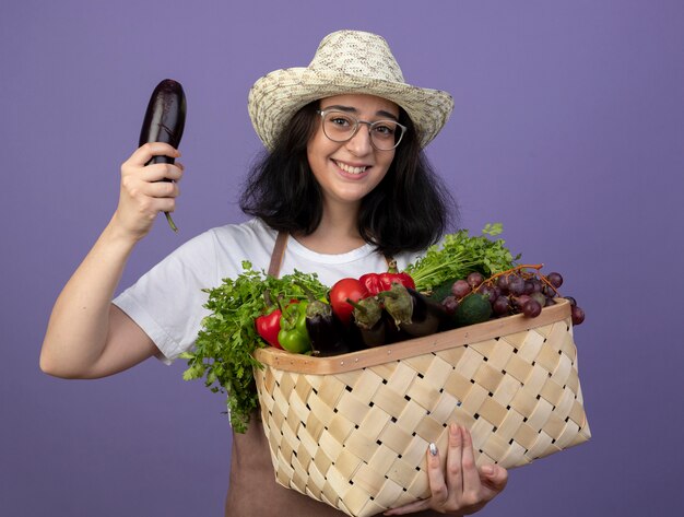 Smiling young brunette female gardener in optical glasses and in uniform wearing gardening hat holds vegetable basket and eggplant isolated on purple wall
