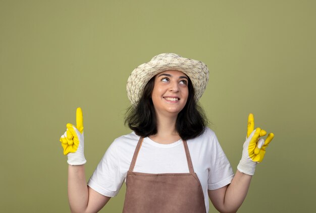 Smiling young brunette female gardener in uniform wearing gardening hat and gloves looks and points up isolated on olive green wall