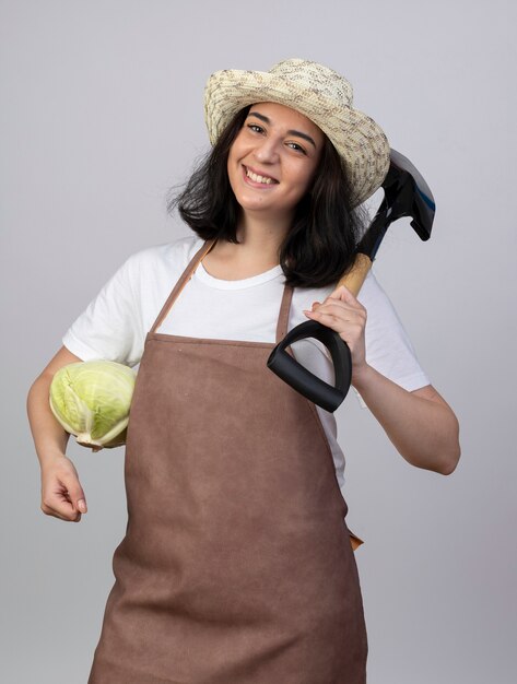 Smiling young brunette female gardener in uniform wearing gardening hat holds cabbage and spade on shoulder isolated on white wall