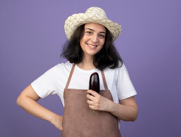 Smiling young brunette female gardener in uniform wearing gardening hat holds eggplant isolated on purple wall