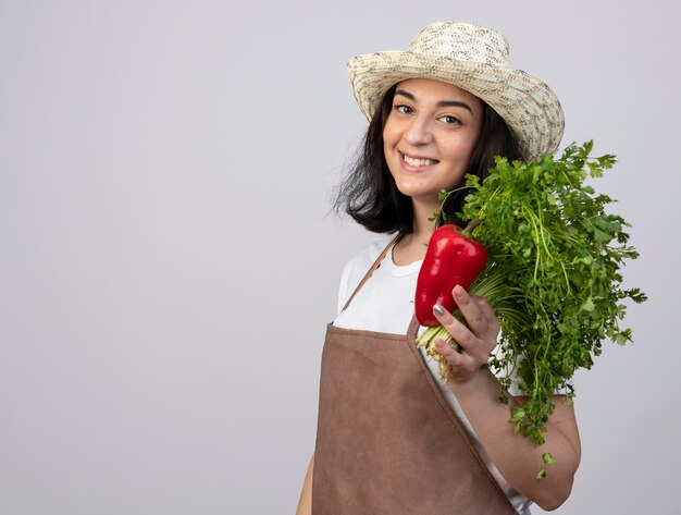 Smiling young brunette female gardener in uniform wearing gardening hat holds red pepper