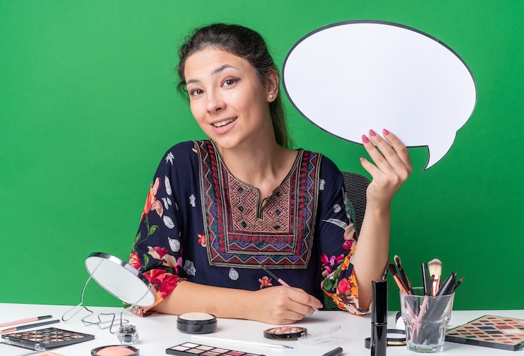 मुफ़्त फोटो smiling young brunette girl sitting at table with makeup tools holding speech bubble