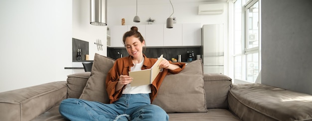 무료 사진 미소 짓는 젊은 갈색 여성은 거실의 소파에 앉아 노트북을 들고 노트 공부를 읽습니다.