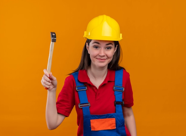 Smiling young builder girl holds hammer and looks at camera on isolated orange background