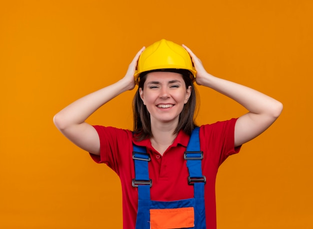 Smiling young builder girl holds head with both hands on isolated orange background