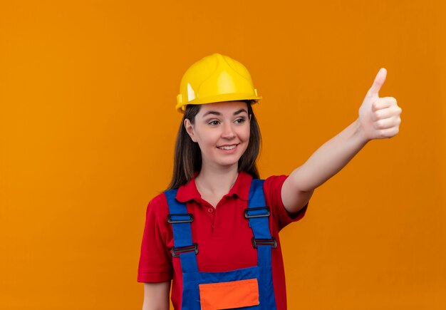 Smiling young builder girl thumbs up and looks to the side on isolated orange background with copy space