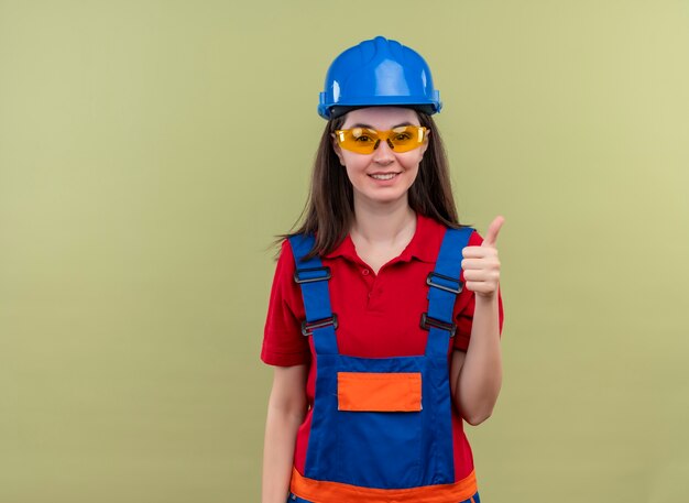 Smiling young builder girl with blue safety helmet and with safety glasses thumbs up on isolated green background with copy space