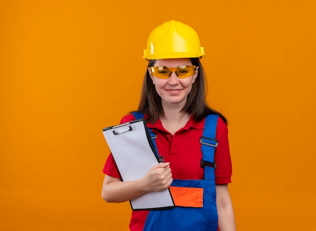 Smiling young builder girl with safety glasses holds clipboard on isolated orange background