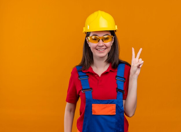 Smiling young builder girl with safety glasses shows victory hand gesture on isolated orange background with copy space