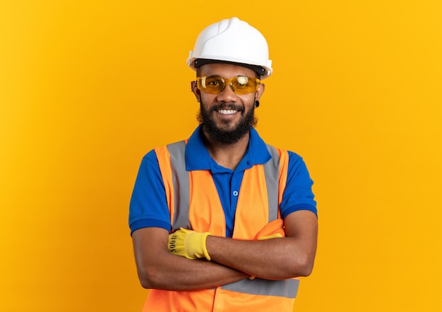 улыбающийся молодой строитель в защитных очках в униформе с защитным шлемом, стоящий со скрещенными руками, изолированными на оранжевой стене с копией пространства