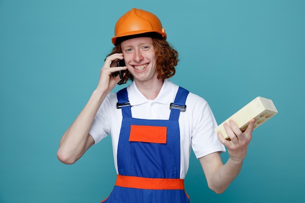 Smiling young builder man in uniform holding brick and speak on the phone isolated on blue background