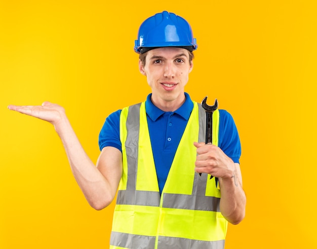 Smiling young builder man in uniform holding open-end wrench and points with hand at side isolated on yellow wall with copy space