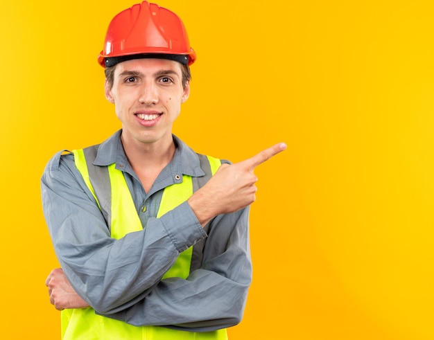 Smiling young builder man in uniform points at side isolated on yellow wall with copy space