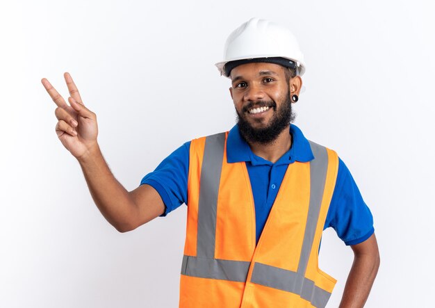 smiling young builder man in uniform with safety helmet gesturing victory sign isolated on white wall with copy space