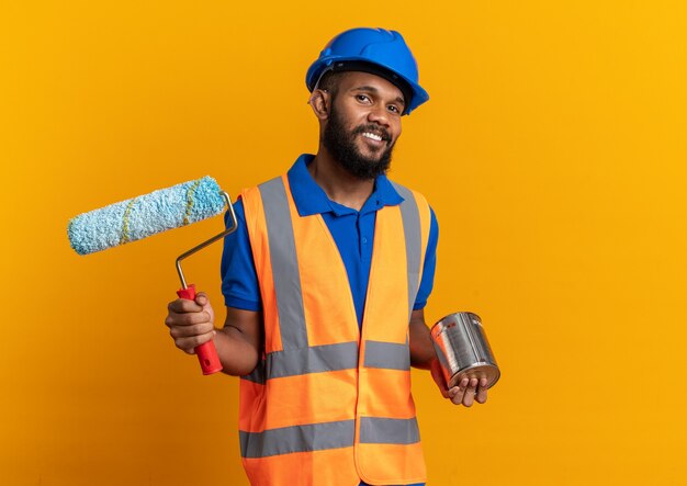 smiling young builder man in uniform with safety helmet holding oil paint and paint roller isolated on orange wall with copy space