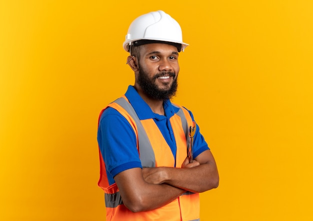 복사 공간이 있는 주황색 벽에 팔짱을 끼고 서 있는 안전 헬멧을 쓴 제복을 입은 웃고 있는 젊은 건축업자