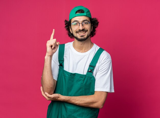 Smiling young builder man wearing uniform with cap points at up 