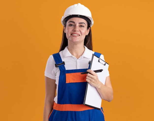 Улыбающаяся молодая женщина-строитель в униформе, держащая буфер обмена с ручкой, изолированной на оранжевой стене