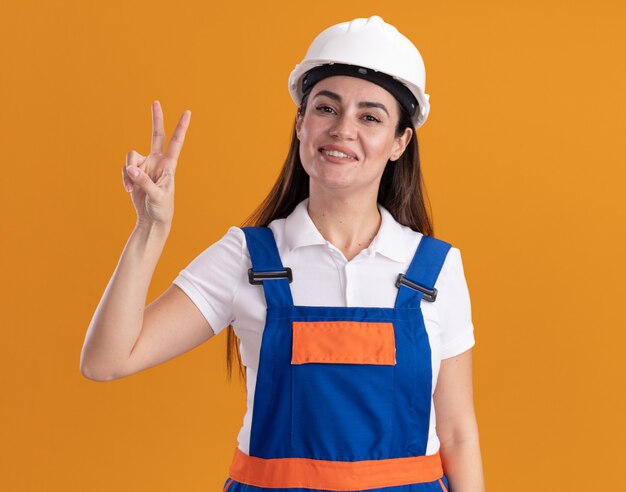 Улыбающаяся молодая женщина-строитель в униформе, показывающая жест мира, изолирована на оранжевой стене