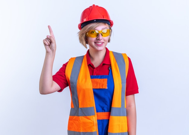 Улыбающаяся молодая женщина-строитель в униформе с очками указывает на вверх изолированной на белой стене