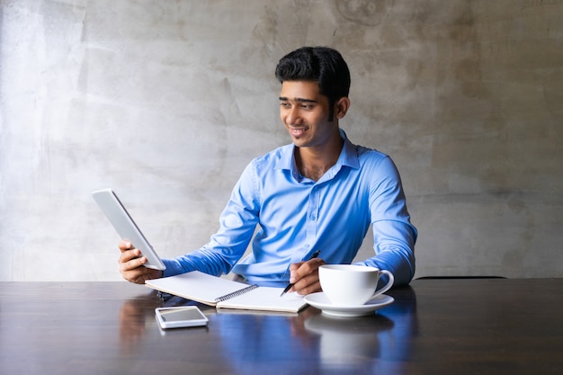 Free photo smiling young businessman drinking coffee at cafe, having video call and writing notes.