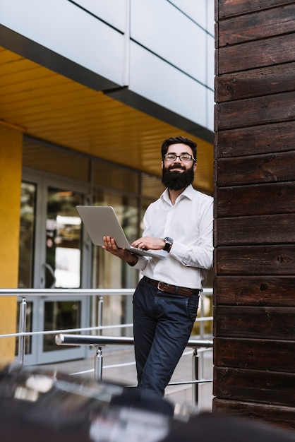 Smiling young businessman holding laptop in hand leaning on wall