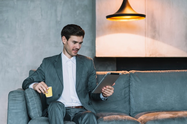 Smiling young businessman sitting on sofa holding coffee cup and digital tablet