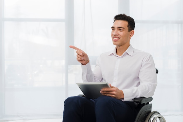 Smiling young businessman sitting on wheelchair holding digital tablet in hand pointing his finger to side