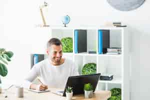 Free photo smiling young businessman using laptop while writing on notepad