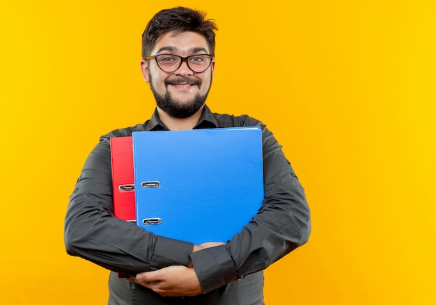 Smiling young businessman wearing glasses holding folders isolated on yellow