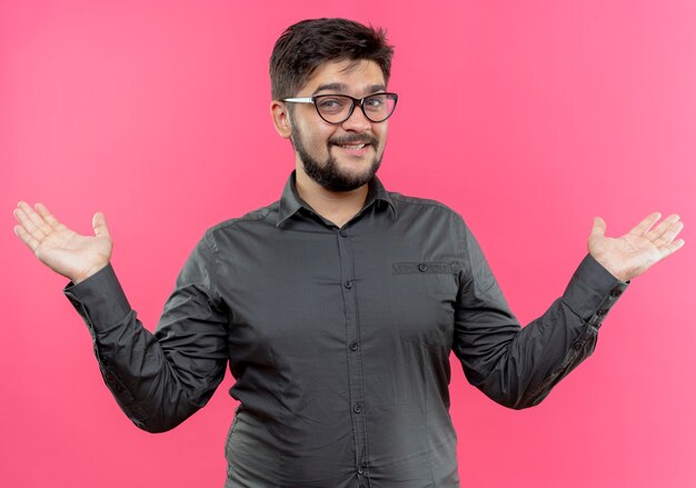 Smiling young businessman wearing glasses spreads hands isolated on pink