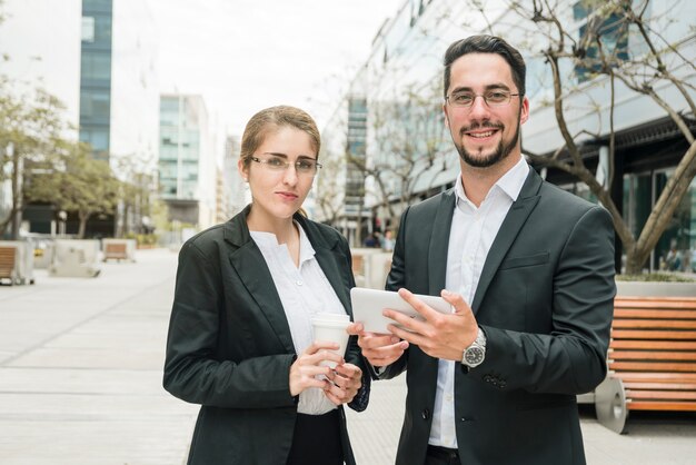 Smiling young businesswoman and businessman standing outside the office holding mobile and disposable coffee cup