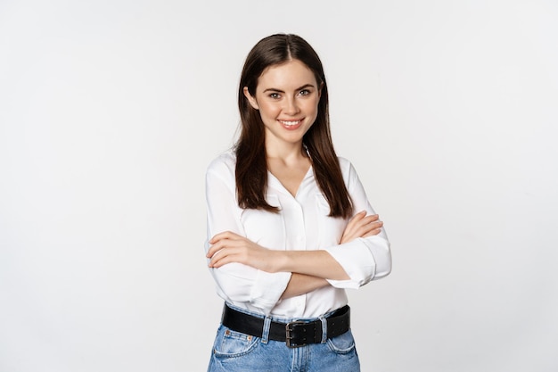 Smiling young businesswoman, female entrepreneur in white shirt, cross arms on chest like professional, standing over studio background.