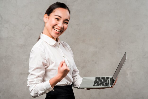 Smiling young businesswoman holding laptop in hand clenching her fist against grey wall
