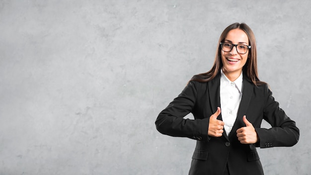 Smiling young businesswoman showing thumb up sign against gray backdrop