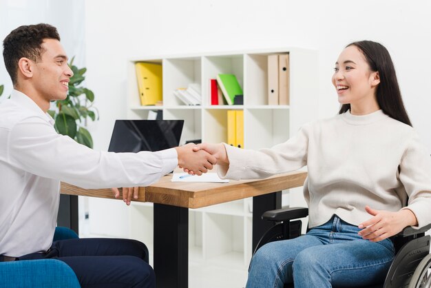 Smiling young businesswoman sitting on wheelchair shaking hands with businessman in the office