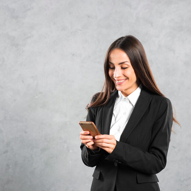 Smiling young businesswoman using smart phone standing against gray concrete backdrop