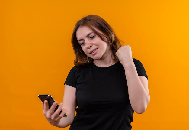 Улыбающаяся молодая случайная женщина держит мобильный телефон и смотрит на него с поднятым кулаком на изолированном оранжевом пространстве