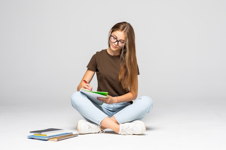 Free Photo smiling young casual woman writing on her notepad while sitting on white wall