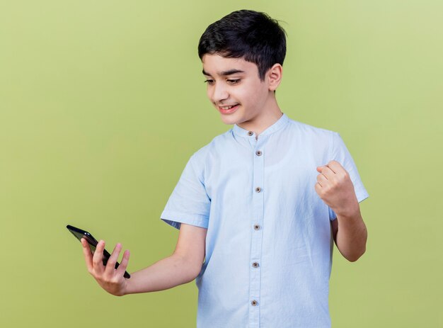 Smiling young caucasian boy holding and looking at mobile phone doing yes gesture isolated on olive green background