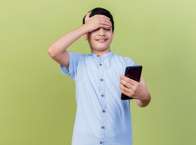 Smiling young caucasian boy holding and looking at mobile phone keeping hand on forehead isolated on olive green background with copy space