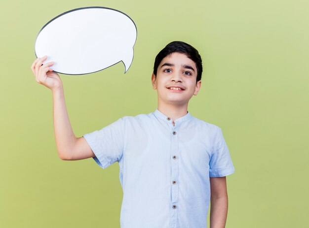 Smiling young caucasian boy holding speech bubble looking at camera isolated on olive green background with copy space