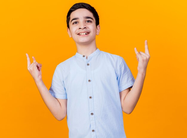 Smiling young caucasian boy looking at camera doing rock sign isolated on orange background
