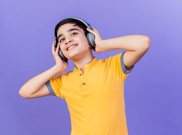 Smiling young caucasian boy looking at camera touching headphones isolated on purple background