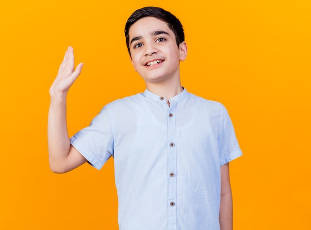 Smiling young caucasian boy looking at camera waving isolated on orange background with copy space