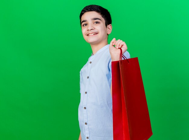 Smiling young caucasian boy standing in profile view holding shopping bag looking at camera isolated on green background with copy space