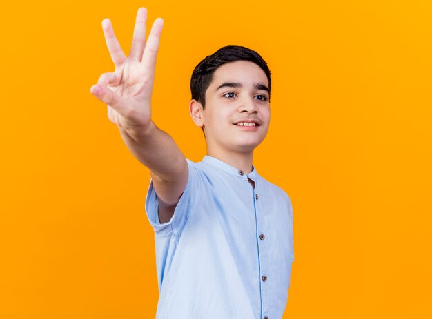복사 공간 오렌지 배경에 고립 된 측면을보고 손으로 3을 보여주는 프로필보기에 서있는 어린 백인 소년 미소
