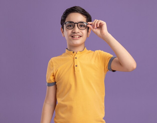 Smiling young caucasian boy wearing and grabbing glasses  isolated on purple wall with copy space