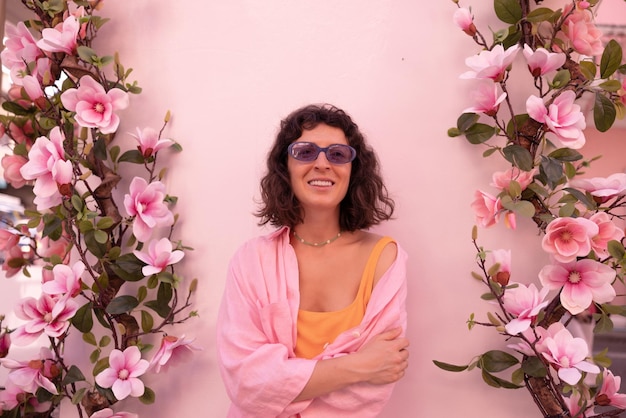 Smiling young caucasian brunette girl in pink casual clothes and glasses with her arms crossed on background of flower wall