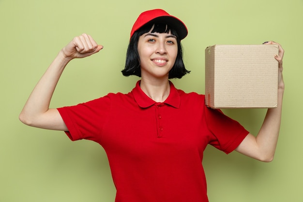 Smiling young caucasian delivery girl holding cardboard box and tensing her biceps